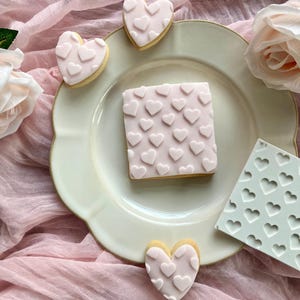 May include: A collection of heart-shaped and square cookies with pink icing and heart-shaped decorations. The cookies are arranged on a white plate with a pink cloth background, alongside a heart-shaped mold.