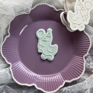May include: A light purple scalloped plate with a white rim holds a light green goose-shaped cookie cutter and a cookie. The cookie cutter and cookie feature a goose wearing a party hat and holding a balloon.