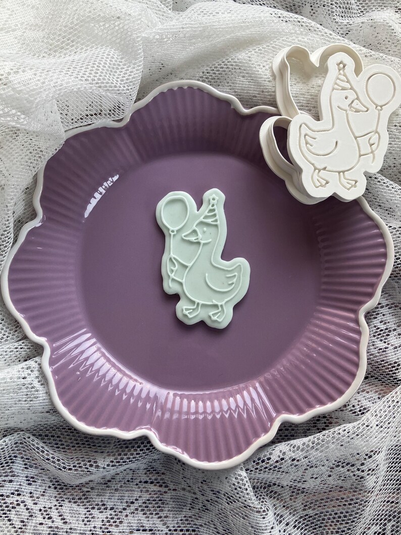 May include: A light purple scalloped plate with a white rim holds a light green goose-shaped cookie cutter and a cookie. The cookie cutter and cookie feature a goose wearing a party hat and holding a balloon.