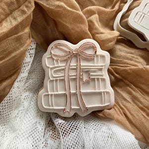 May include: A white cookie cutter in the shape of a stack of books tied with a bow. The bow and book details are outlined in copper. The cutter sits on a tan and white fabric background.