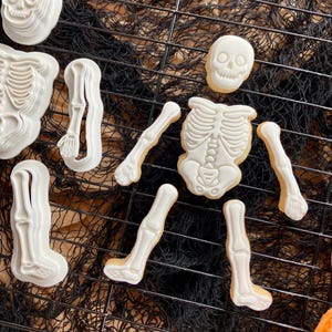 May include: A selection of Halloween skeleton cookies and matching cookie cutters. The cookies are white with detailed bone designs, and the cutters are also white. The cookies are arranged on a black wire rack over an orange surface.