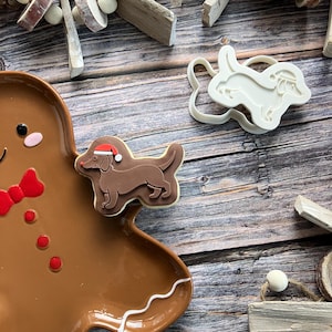May include: A brown gingerbread man-shaped plate with a dachshund cookie decorated with a Santa hat. A white dachshund cookie cutter is also present. The scene is set on a wooden surface with decorative wooden elements and pinecones.