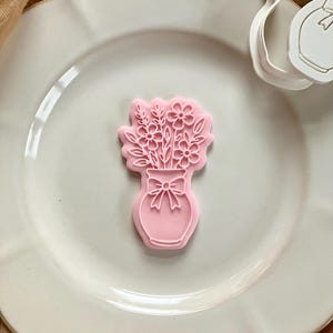 May include: A pink cookie cutter in the shape of a vase filled with flowers and a bow. The cutter is on a white plate with a gold-colored cloth in the background. The cutter is made of plastic.