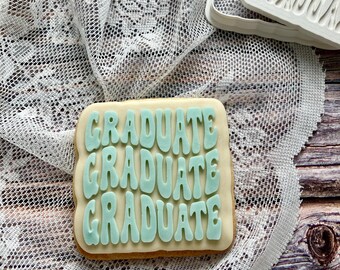 May include: A square cookie with the word "GRADUATE" in light blue icing. The word is repeated three times. The cookie sits on a white lace cloth. A white plastic cookie cutter with the word "GRADUATE" is in the background.