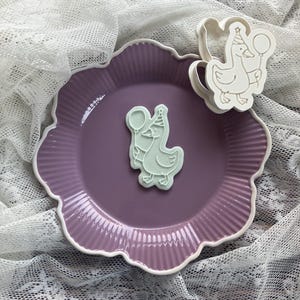 May include: A light purple ceramic plate with a white scalloped edge, holding a light green goose-shaped cookie cutter and a cookie. The goose holds a balloon and wears a party hat. The plate sits on a white lace cloth.