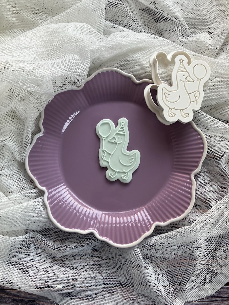 May include: A light purple ceramic plate with a white scalloped edge, holding a light green goose-shaped cookie cutter and a cookie. The goose holds a balloon and wears a party hat. The plate sits on a white lace cloth.