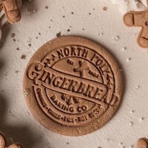 May include: A circular gingerbread cookie, embossed with the words "North Folk Gingerbread Baking Co.", sits on a frosted surface. Other gingerbread biscuits and decorations surround it.