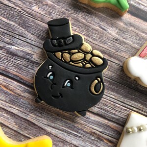 May include: A black cookie shaped like a pot of gold with a top hat, filled with gold coins. The cookie has a smiling face with blue eyes and is decorated with black icing. Other cookies are visible in the background.