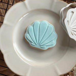 May include: A light blue seashell-shaped biscuit on a white, scalloped plate. The biscuit has a detailed fan design. A white seashell cookie cutter is next to the plate. The background includes a brown surface and a black wire rack.