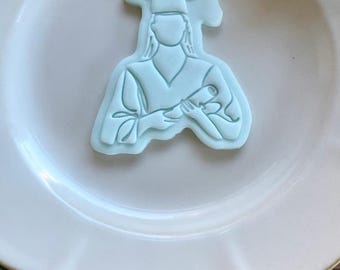 May include: A light blue graduation-themed cookie cutter in the shape of a person wearing a graduation cap and gown, holding a diploma. The cutter is on a white plate, with a white floral-shaped mold in the background.