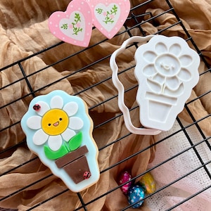 May include: A decorated cookie shaped like a flower in a pot with a light blue background, a white daisy with a yellow center, and two small ladybugs. Beside it is a white plastic cookie cutter and two pink heart-shaped cookies with floral designs.