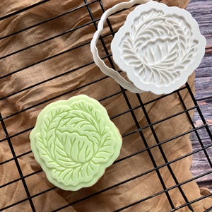 May include: A light green, leaf-shaped biscuit sits on a black wire rack next to a white biscuit cutter. The biscuit cutter is also leaf-shaped. The background is a tan cloth.