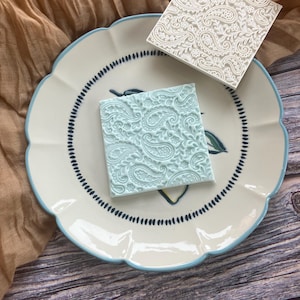 May include: Two square stamps with a paisley pattern, one light blue and one white, are displayed on a scalloped plate with a blue rim and dotted border. The plate rests on a wooden surface, with a tan cloth behind.