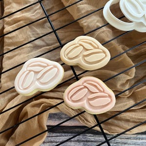May include: Three coffee bean-shaped cookies in varying shades of beige and pink, arranged on a black wire cooling rack. A white cookie cutter is also visible. The cookies have a detailed design of coffee beans.