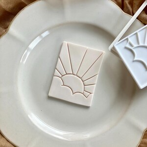 May include: A rectangular off-white clay cutter featuring a debossed sun rising over clouds design. The tool has a flat surface with recessed lines forming the sun rays and cloud outlines, positioned next to a matching white plastic cutter frame.