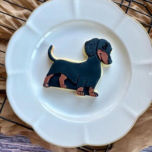 May include: A dachshund-shaped cookie with black, brown, and tan icing sits on a white, scalloped-edge plate. The cookie is placed on a metal cooling rack over a tan cloth.