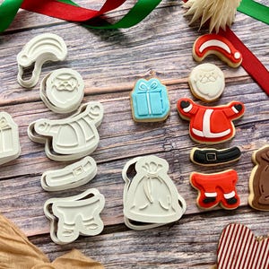 May include: A selection of Christmas-themed cookie cutters and decorated biscuits. The cutters are white plastic, shaped like Santa Claus, gifts, and clothing. The biscuits are decorated with red, white, blue, and brown icing.