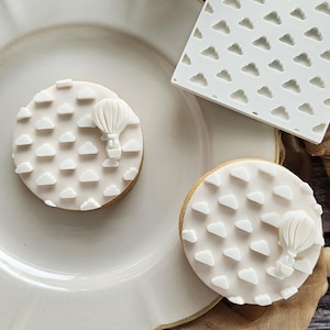 May include: Two round cookies topped with smooth off-white icing featuring a raised pattern of small clouds and a single hot air balloon. A square white plastic texture mat with a repeating cloud pattern is positioned next to the cookies.