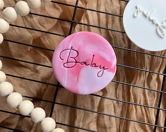 May include: A pink and white marbled round cookie with the word "Baby" in cursive script. A white round cookie with cursive script is to the right. The cookies are on a black wire rack with a wooden bead garland and a tassel.