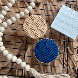 May include: Three cookies with a plaid design, one beige, one blue, and one on a white square. The cookies are on a black wire rack, with a wooden bead garland and a tassel in the foreground. The background is a tan fabric.