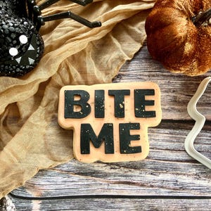 May include: A Halloween-themed image featuring a cookie with the words "BITE ME" in black icing. The cookie is surrounded by a black spider, a plush pumpkin, and a decorative cloth on a wooden surface.