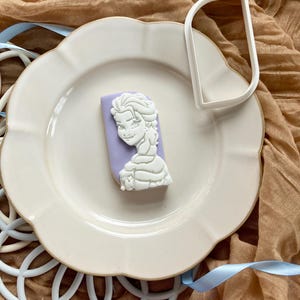 May include: A white cookie with a princess design on a light purple background, placed on a white plate. A white cookie cutter is next to the cookie. The plate is on a brown cloth with a blue ribbon.
