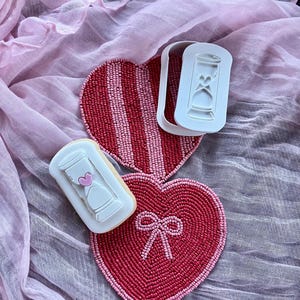 May include: A Valentine's Day-themed image featuring heart-shaped beaded coasters in red and pink, along with a white cookie cutter and a decorated cookie with an hourglass design. The background is a pink fabric.