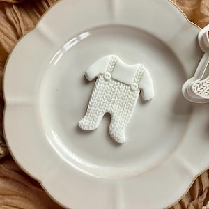 May include: A white baby onesie-shaped cookie cutter with a textured knit pattern. The onesie has small straps and buttons. The cutter is on a white plate with a scalloped edge, next to a second cutter.