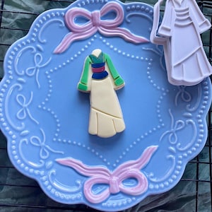 May include: A light blue, scalloped-edge plate with pink bows and a cookie shaped like a traditional dress. A white plastic dress-shaped cookie cutter is also present. The cookie has green, blue, and pink accents.