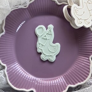 May include: A light green cookie cutter in the shape of a goose wearing a party hat and holding a balloon. The cookie cutter is on a purple plate with a white scalloped edge. Another cookie cutter is visible.