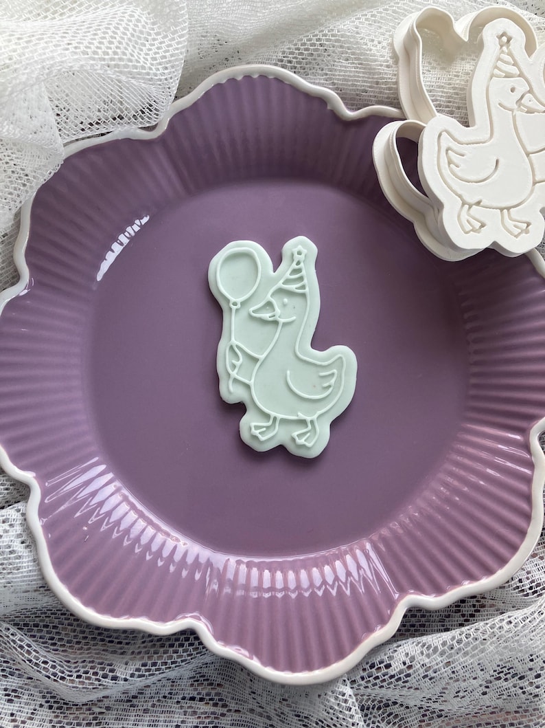 May include: A light green cookie cutter in the shape of a goose wearing a party hat and holding a balloon. The cookie cutter is on a purple plate with a white scalloped edge. Another cookie cutter is visible.
