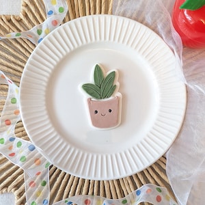May include: A sugar cookie shaped like a potted plant with green leaves and a light pink pot featuring a simple black-lined face with two circular eyes and a small curved smile, centered on a white plate with a ribbed edge design.