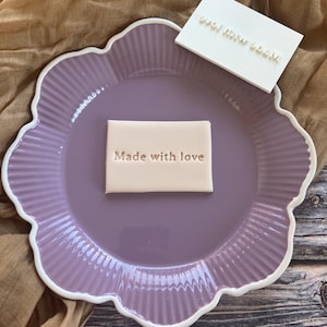 May include: A lavender scalloped plate with a white rim holds two rectangular stamps. One stamp is light beige with the words "Made with love" embossed. The other stamp is white with the same text.