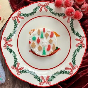 May include: A white ceramic plate with a red rim and a festive design. The plate features a garland of green leaves and red bows around the edge. A decorated cookie with Christmas ornaments sits in the center. Red berries are in the background.