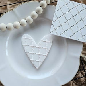 May include: A white heart-shaped cookie with a lattice design sits on a white plate. A white square with a similar lattice pattern is next to the plate. A string of wooden beads is above the plate, and the background is a tan fabric.