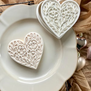 May include: A heart-shaped cookie with a floral design, next to a white plastic cookie cutter with the same pattern. The cookie is light brown with a detailed embossed floral pattern. The cutter is white and has a matching floral design. The items are on a white plate.