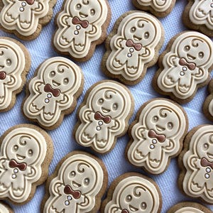 May include: A close-up shot of several gingerbread man biscuits. Each biscuit has a light brown base with a lighter tan icing in the shape of a gingerbread man. The gingerbread men have brown bow ties and smiling faces.