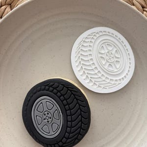 May include: A black and grey tire-shaped biscuit and a white tire-shaped cookie cutter on a beige plate. The biscuit has a detailed tyre tread and a grey wheel. The cookie cutter is white and has a tyre tread design.