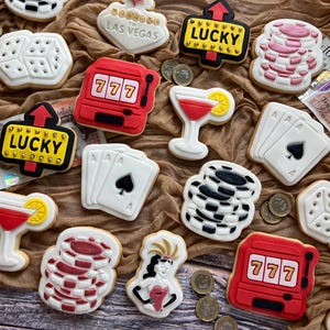 May include: A collection of casino-themed biscuits, including dice, playing cards, slot machines, and cocktail designs. The biscuits are decorated with red, white, and black icing, and some feature the text "LUCKY" and "LAS VEGAS".