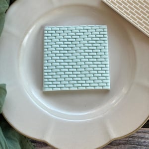 May include: A square, pale blue brick pattern sits on a white plate. The brick pattern is a solid block of rectangular shapes. A white brick pattern is in the upper right corner. The plate has a scalloped edge.