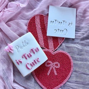 May include: A square cookie with the text "Pickle is TuTu Cute" in pink frosting, next to a heart-shaped beaded coaster and a white stamp with the same text. The coaster is red and pink, and the stamp is white.