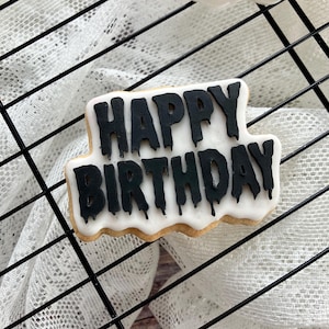 May include: A decorated sugar cookie with the words "HAPPY BIRTHDAY" in black icing. The cookie has a white icing base and a light brown edge. The cookie is on a black wire rack.