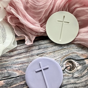 May include: Two round, embossed cross designs in the image. One is a light lavender color, and the other is a pale green. The crosses are simple, with a vertical line intersected by a horizontal line. The background includes pink fabric and a wooden surface.