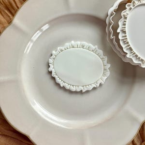 May include: A white ceramic plate with a scalloped edge, holding a white oval-shaped cookie cutter with a ruffled border. Another similar cutter is partially visible. The plate and cutters are on a textured, light brown surface.