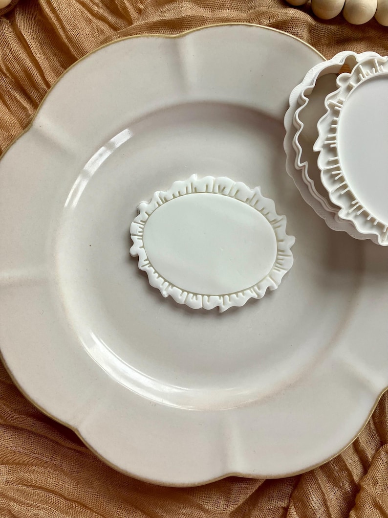 May include: A white ceramic plate with a scalloped edge, holding a white oval-shaped cookie cutter with a ruffled border. Another similar cutter is partially visible. The plate and cutters are on a textured, light brown surface.