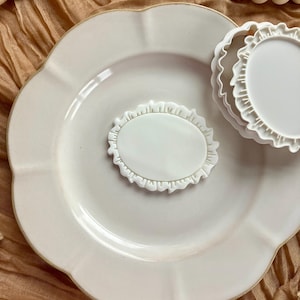 May include: A white ceramic plate with a scalloped edge holds a white oval-shaped cookie cutter with a ruffled border. Another round cookie cutter with a ruffled edge is also present. The background is a tan fabric.