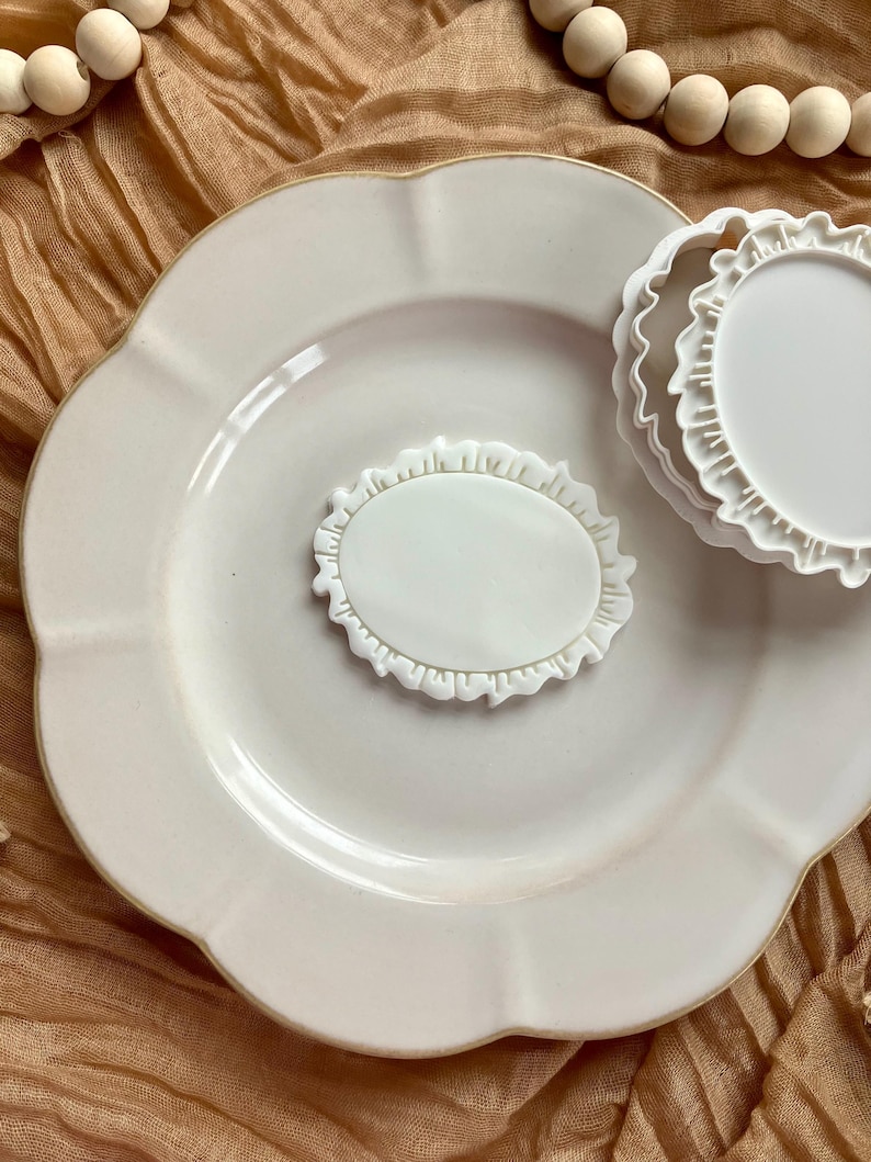 May include: A white ceramic plate with a scalloped edge holds a white oval-shaped cookie cutter with a ruffled border. Another round cookie cutter with a ruffled edge is also present. The background is a tan fabric.