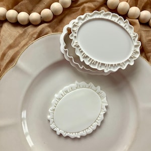 May include: Three white, oval-shaped cookie cutters with a ruffled edge are stacked on a white plate. One cutter is placed separately. A string of wooden beads and a tan cloth are in the background.