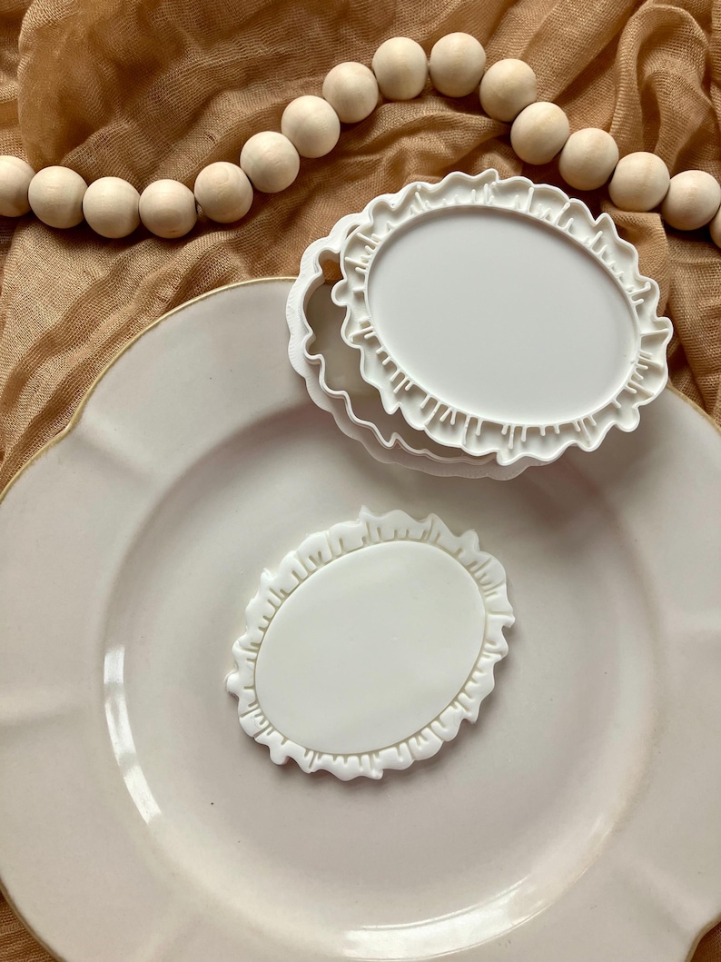 May include: Three white, oval-shaped cookie cutters with a ruffled edge are stacked on a white plate. One cutter is placed separately. A string of wooden beads and a tan cloth are in the background.