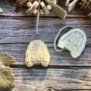 May include: A cookie and a silicone mold, both shaped like a jacket, sit on a wooden surface. The cookie is beige with brown details, while the mold is white. Decorative elements like pinecones and wooden pieces surround the jacket.